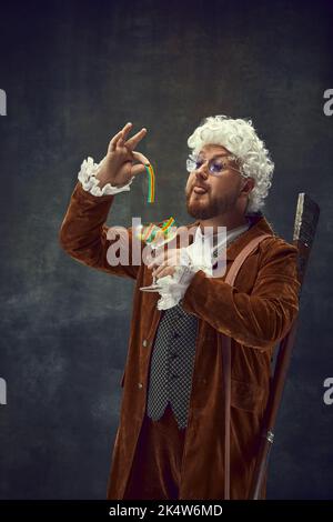 Süßigkeiten. Vintage-Porträt eines jungen Mannes in braunem Vintage-Anzug und weißer Perücke wie mittelalterliche königliche Jäger isoliert auf dunklem Hintergrund. Stockfoto