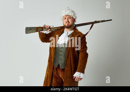 Ernsthafter junger Mann im Bild einer mittelalterlichen Person im Vintage-braunen Jagdanzug und weißer Perücke mit altem Jagdgewehr isoliert auf grauem Hintergrund. Stockfoto