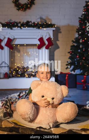 Kind lächelt und umarmt weiches Spielzeug in der Nähe verschwommener Kamin und weihnachtsbaum zu Hause am Abend, Stock Bild Stockfoto