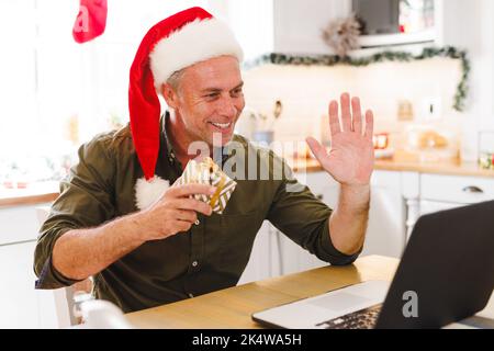Glücklicher kaukasischer Mann mit Weihnachtsmütze, der in der Küche am Tisch saß und einen Laptop für einen Videoanruf benutzte Stockfoto