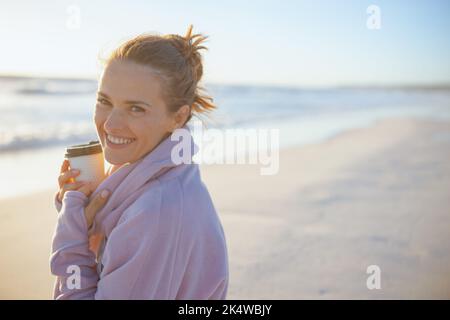 Porträt einer lächelnden eleganten Frau in kuscheligem Pullover mit einer Tasse Heißgetränk am Strand am Abend. Stockfoto