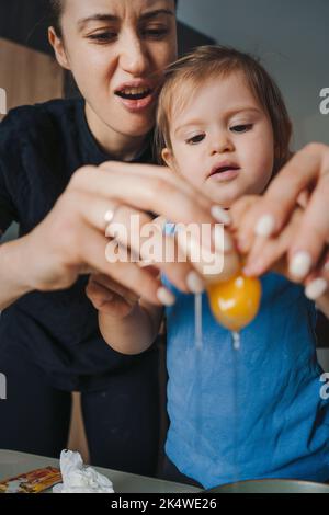 Kaukasische Mutter und ihre Tochter zerbrechen Eier, während sie in der Küche zusammen kochen. Teig zum Backen von Keksen vorbereiten. Lustige Familie. Glücklich Stockfoto