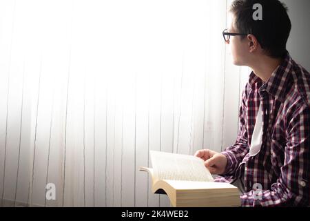 Nahaufnahme eines Mannes mit mittlerem Erwachsenen, der auf einem Stuhl neben einem Fenster sitzt und ein Buch liest Stockfoto