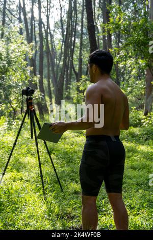 Reifer Sportler mittleren Alters, der in sportlicher Kleidung auf einer Fitnessmatte in Standposition steht, sich online ein Videotutorial zum Training ansieht und vloggt Stockfoto