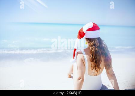 Von hinten gesehen Frau in weißem Badeanzug mit langen welligen Haaren und gestreiftem weihnachtshut am Strand. Stockfoto