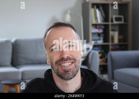 Mann, der zu Hause lächelt. Bärtiger Mann mit lässigem schwarzem Hoodie. Erwachsener kaukasischer Mann im Jahr 30s oder 40s lächelte in Richtung Kamera. Stockfoto