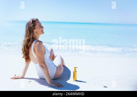 Entspannte, stylische Frau mit spf in weißem Badeanzug am Strand. Stockfoto