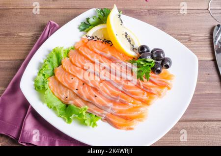 Frischer roter Fisch, der auf einem weißen Teller auf dunklem Holzgrund in Streifen mit Zitrone und Kräutern geschnitten wurde Stockfoto