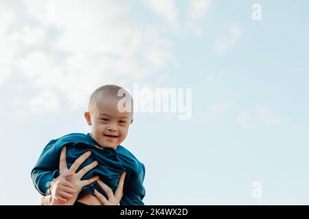 Niedlicher blonder Junge mit Down-Syndrom auf den Armen der Mutter, blauer Himmel Hintergrund Stockfoto