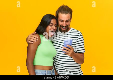 Glückliches multirassisches Paar überrascht Blick auf ein Mobiltelefon, isoliert über gelbem Hintergrund. Stockfoto