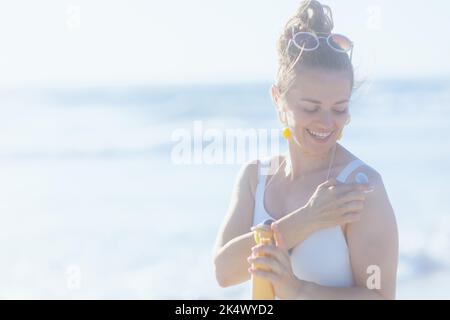 Lächelnde, moderne Frau in weißem Badeanzug am Strand, die spf anwendet. Stockfoto