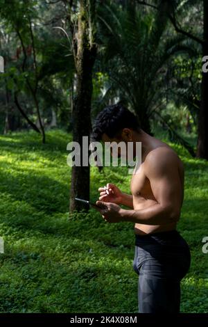 Aktive, gutaussehende männliche Yogi spazieren in einem Park durch den Wald vor oder nach dem Training. Yoga-Kurs für junge hispanische Sportler mit einer Fitnessmatte Stockfoto