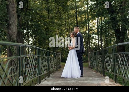 Im Sommer steht ein tolles Hochzeitspaar im Park. Junge hübsche Frau mit Blumenstrauß, Bräutigam in Anzug umarmt Braut. Stockfoto