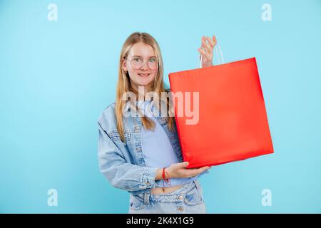 Porträt eines gut aussehenden Teenagers mit einem riesigen roten Papierpaket auf blauem Hintergrund. Einkaufen, Verkauf, Studio. Stockfoto