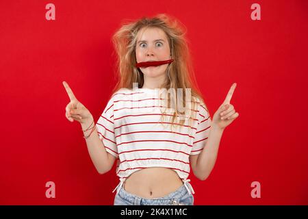 Porträt eines erstaunten Teenagers mit zerzausten, schönen Haaren, die die Hände mit den Fingern nach oben heben und im Mund rote Chilli halten. Stockfoto