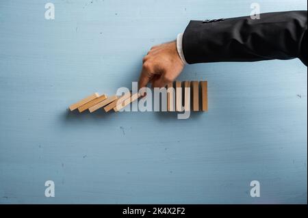 Hand eines Geschäftsmannes, der die einstürzenden Dominos in einem konzeptuellen Bild des Krisenmanagements stoppt. Auf pastellblauem Holzhintergrund. Stockfoto