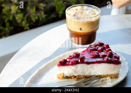 Eine einzelne Scheibe oder Portion frisch zubereiteter, nachgiebiger Kirschkäsekuchen. Es wird auf einem Teller mit einem Eiskaffee an einem Außentisch in einem Café serviert. Stockfoto