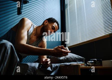 Junger, schlafloser Mann, der nachts auf dem Bett sitzt und eine Flasche Schlaftabletten in der Hand anschaut, während er eine einschläft Stockfoto