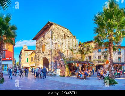 SIRMIONE, ITALIEN - 10. APRIL 2022: Panorama der überfüllten Touristenstraße Via Flaminia mit kleinen Restaurants im Freien, historischen Häusern, hohen Palmen und Stockfoto