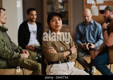 Junge gestresste hispanische Frau mit gekreuzten Armen auf der Brust und Blick auf die Kamera, während sie gegen interkulturelle Männer sitzt, die diskutieren Stockfoto