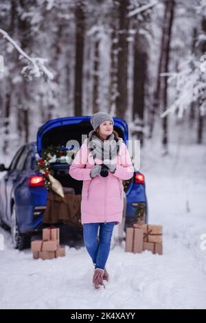 Eine Frau in einem winterverschneiten Wald im Kofferraum eines Autos, das mit Weihnachtsdekor dekoriert ist. Eine Fotografin hält eine Kamera in den Händen. Stockfoto