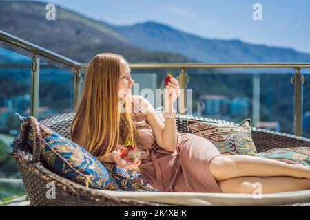 Portrait einer wunderschönen Frau in einem wunderschönen Kleid, die auf einer Terrasse sitzt und Erdbeere isst Stockfoto