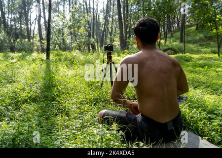 Junger Mann, der Yoga Asana praktiziert, balanciert, meditiert, während er auf einem Bein auf einer Sportmatte auf grünem Gras im Park steht. Tablet für Online-Kurs verwenden. Stockfoto