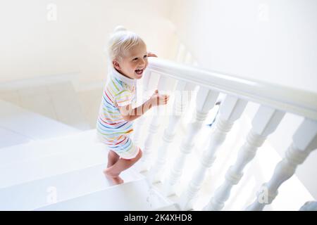 Kind geht Treppe im weißen Haus. Kleiner Junge, der im sonnigen Treppenhaus spielt. Familie zieht in ein neues Zuhause. Kinder kriechen Stufen der modernen Treppe. Stockfoto