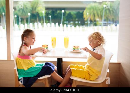 Kinder essen Regenbogenkuchen im Restaurant. Junge und Mädchen mit Süßigkeiten und Gebäck. Kinder trinken frischen Orangensaft im Café. Familie, die im Stadtcafé auswärts isst Stockfoto