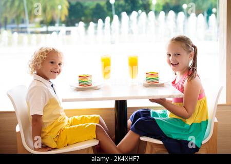 Kinder essen Regenbogenkuchen im Restaurant. Junge und Mädchen mit Süßigkeiten und Gebäck. Kinder trinken frischen Orangensaft im Café. Familie, die im Stadtcafé auswärts isst Stockfoto