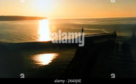 Whitby, North Yorkshire, Großbritannien, liegt zwar an der Ostküste, aber tatsächlich nach Norden, was zur Tatsache führt, dass es eine von nur zwei britischen Städten ist, in denen die Sonne im Meer untergeht. Sie ist besonders zur Sommersonnenwende prominent. Die Aussicht zeigt den Sonnenuntergang vom Kettleness Point aus, mit Blick auf den St. Mary's Kirchhof Stockfoto