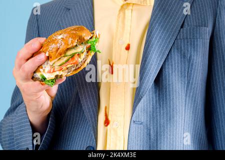 Die Hand eines Mannes hält einen Hamburger. Schmutzige Ketchup-Flecken auf einem gelben Hemd. Stockfoto