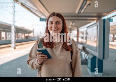 Junge Rotschopf-Frau wartet auf Bahnsteig mit Rucksack und Pass. Konzept des Eisenbahnverkehrs, Reisender. Stockfoto