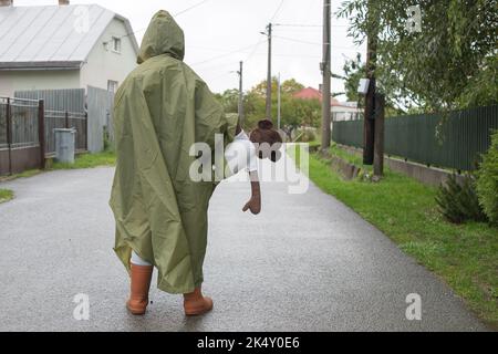 Junge mit grünem Regenmantel, der an regnerischen Tagen auf der Straße steht. Kinderportrait im Herbst kaltes Wetter. Hält weiches Affen Spielzeug. Platz für Text kopieren. Stockfoto