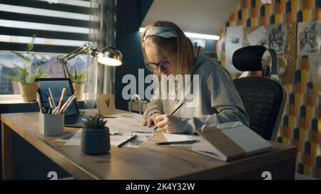 Eine blonde Frau, die an einem Storyboard in einem heimischen Designstudio arbeitet. Fokussierte Frau in einem Kapuzenpullover mit Bleistiftskizzen. Das Storyboard als Roadmap für das Video. Stockfoto