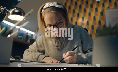 Die fokussierte Illustratorin zeichnet Skizzen für ihr Projekt. Die junge Künstlerin mit einer Brille, die in einem grauen Hoodie gekleidet ist, sitzt auf ihrem Schreibtisch. Macht Bleistiftskizzen. Konzept zum Erzählen von Geschichten. Stockfoto