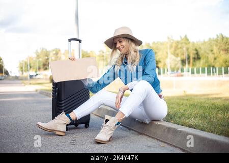 Porträt einer jungen glücklichen Frau, die leere Pappe hält, auf der Straße in der Nähe eines schwarzen Koffers sitzt, grünes Gras. Reisen. Stockfoto