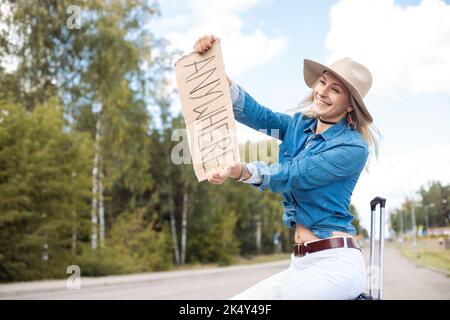 Junge erstaunliche blonde Frau, die Hände mit Pappe mit Inschrift überall, sitzt auf schwarzem Koffer in der Nähe des Waldes. Stockfoto