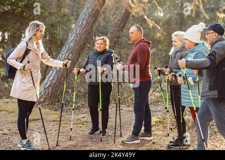 Blonde Trainerin, Sporttrainerin mit einer Gruppe von Menschen lehren skandinavisches Gehen im Wald. Leitender Körperkurs Stockfoto