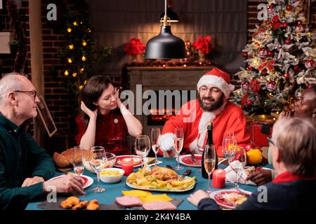 Mann im weihnachtsmann-Kostüm feiert weihnachten mit der Familie und trinkt Sekt zum festlichen Abendessen. Feiertagsfeier, Zusammenkunft der Menschen, traditionelles Essen Stockfoto