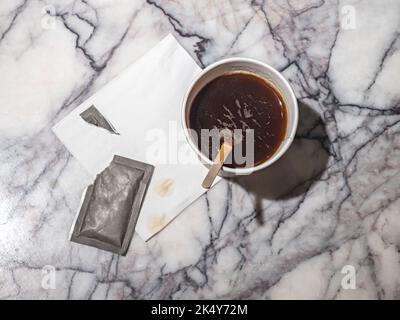 Schwarzer Kaffee in der braunen Einweg-Tasse, ein zerrissener Beutel mit Zucker und der hölzerne Rührstab auf dem Hintergrund der Marmorplatte Stockfoto