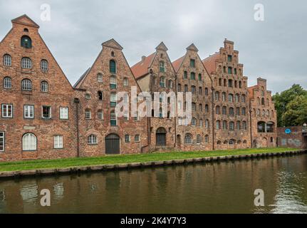 Deutschland, Lübeck - 13. Juli 2022: Reihe von rot-braunen historischen Salzlagerhäusern entlang des Trave-Flusses unter bläulicher Wolkenlandschaft. Grüner Rasen am Kai. Stockfoto