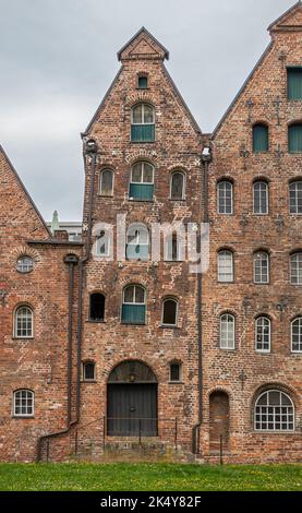 Deutschland, Lübeck - 13. Juli 2022: Kleinste und schräge rotbraune historische Salzlagerhallen entlang des Flusses Trave unter grauer Wolkenlandschaft. Grünes Gesetz Stockfoto