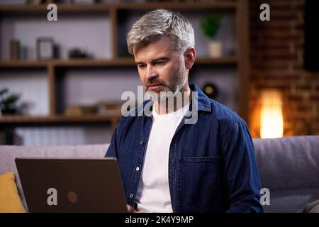 Ein freiberuflicher Mann, der einen Laptop verwendet, arbeitet ferngesteuert und sitzt auf der Couch. Fokussierter Mann, der während der Arbeit an einer Videokonferenz auf einem Laptop teilnimmt Stockfoto
