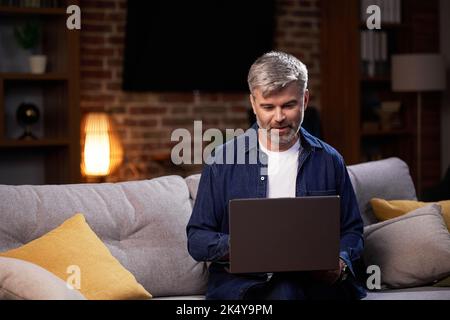Ein freiberuflicher Mann, der einen Laptop verwendet, arbeitet ferngesteuert und sitzt auf der Couch. Fokussierter Mann, der während der Arbeit an einer Videokonferenz auf einem Laptop teilnimmt Stockfoto