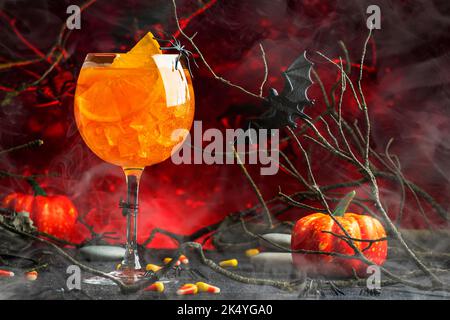 Halloween aperol Spritz Cocktail auf gruselig dunkelblutig rotem Hintergrund mit verdrehten Ästen, Fledermäusen, Steinen und Spinnen, festlicher Drink zur Party Stockfoto