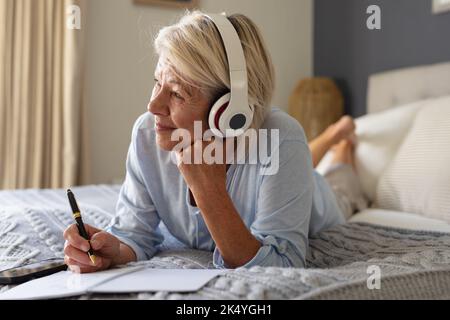 Glückliche ältere kaukasische Frau, die im Schlafzimmer auf dem Bett lag, Notizen gemacht und Kopfhörer getragen hat Stockfoto