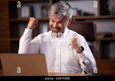 Der glückliche Geschäftsmann war von den guten Nachrichten überrascht und machte eine Ja-Geste, während er auf den Laptop im Büro schaute. Trader genießen Erfolg, feiern Stockfoto