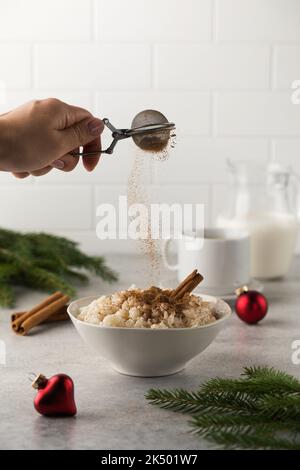 Streuen Sie Zimt über skandinavischen Reisbrei in einer Schüssel, die von Weihnachtskugeln umgeben ist. Frühstück am Silvesterabend. Stockfoto