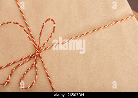 Draufsicht auf ein Weihnachtsgeschenk, eingewickelt in neutralem Papier und gebunden mit einer roten und goldenen Zierschnur. Einfaches Nahaufnahme-Bild mit Kopierplatz. Stockfoto